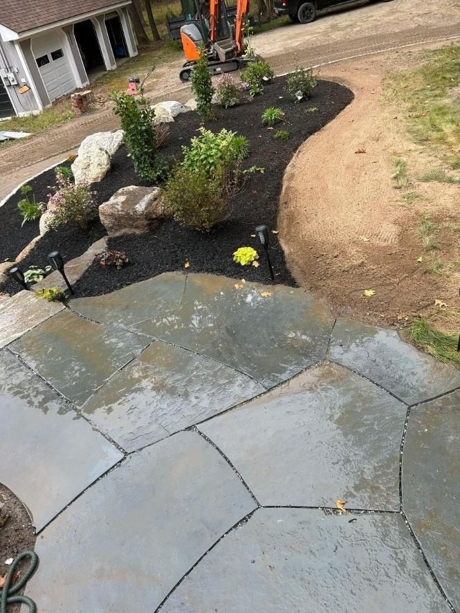 Slate patio beside a freshly landscaped garden bed with rocks, shrubs, and a compact orange excavator