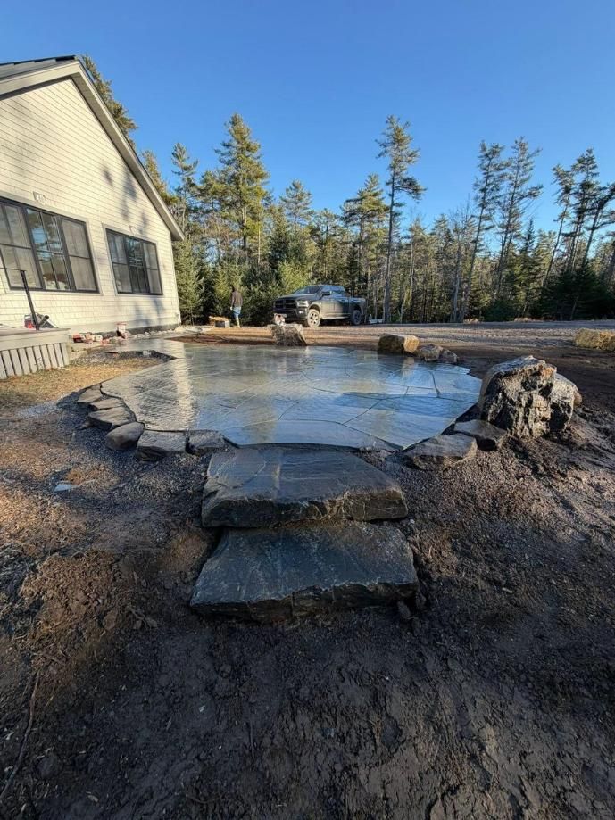 Stone patio and fire pit beside a house, with trees and a clear blue sky in the background.