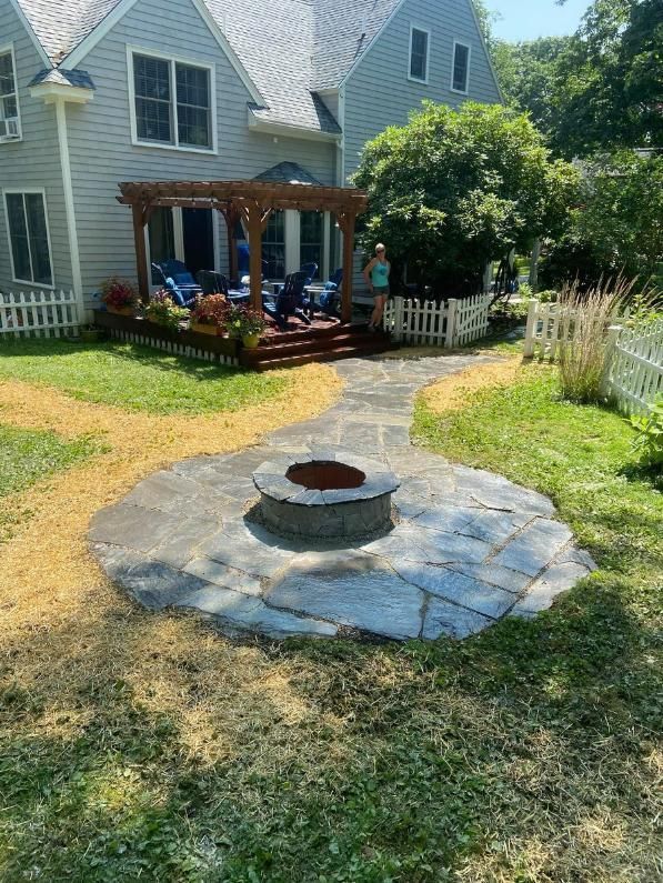Backyard fire pit in a stone circle beside a house with a wooden deck and shaded lawn.