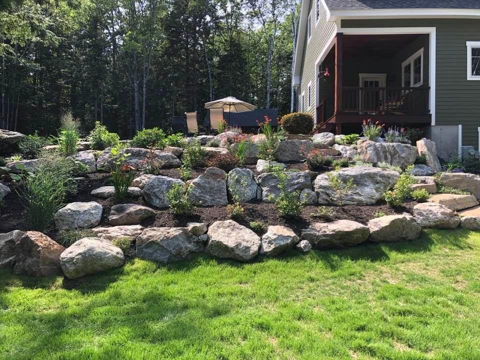 Rock terraced garden beside a house, with lush green lawn and trees in the background.