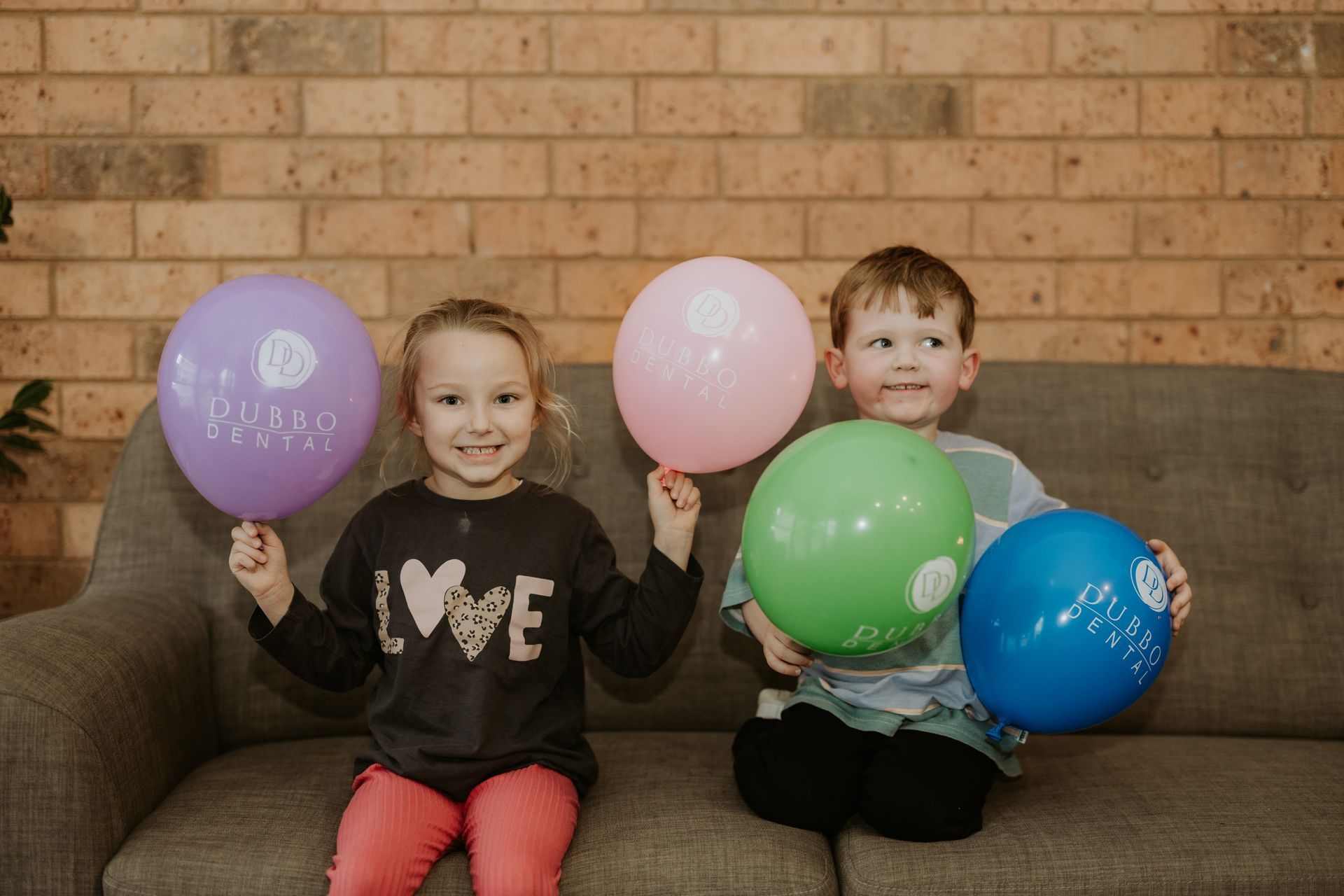 Two Smiling Children Holding Colourful Balloons on a Couch — Dubbo Dental in Dubbo, NSW