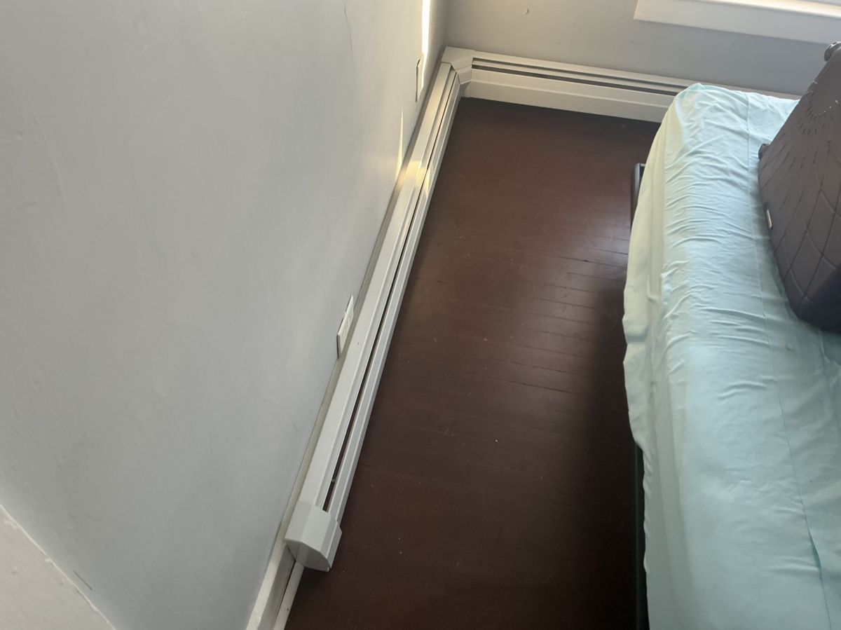 White baseboard heating units run along the corner of a room with dark wood flooring and a bed with blue linens.