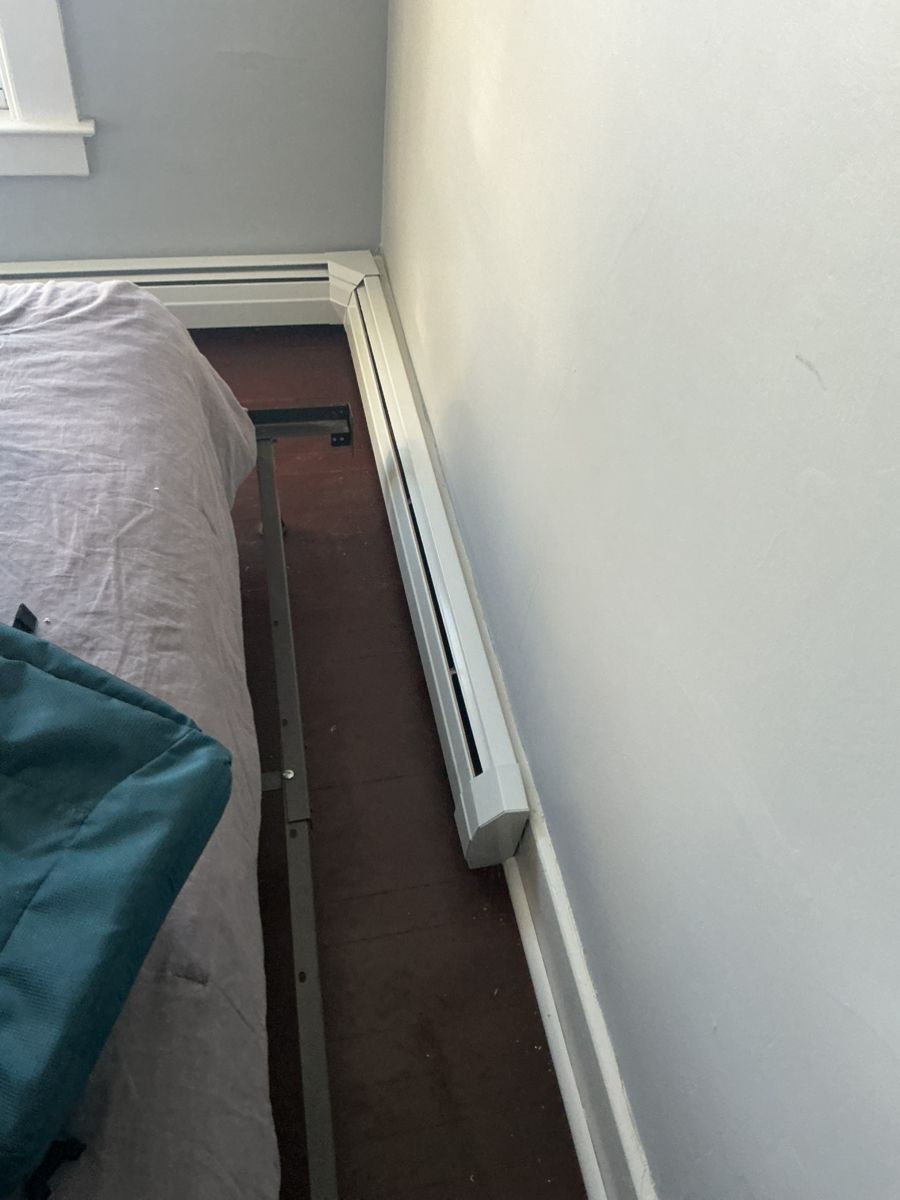 A white baseboard heater runs along a wall near the side of a bed with a dark frame on a carpeted floor.