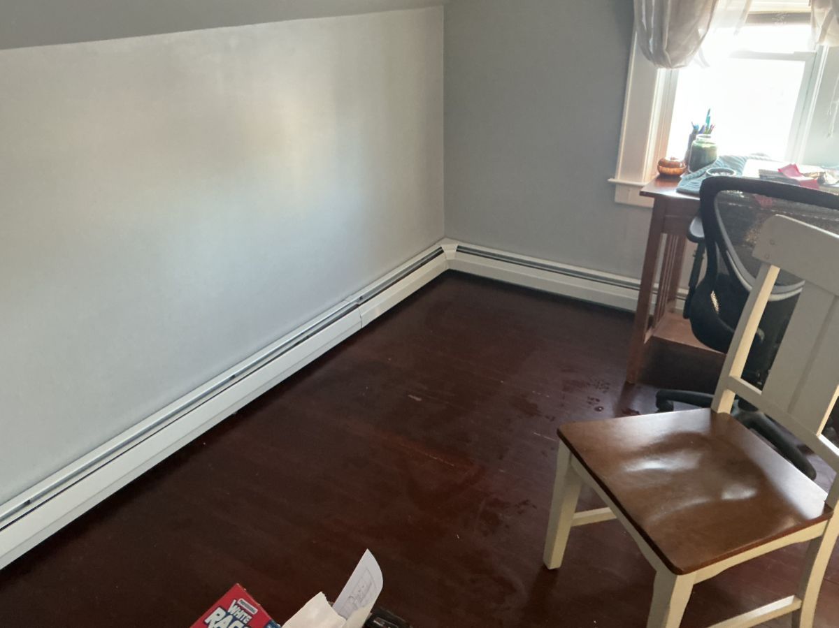 A corner of a room with dark wood flooring, grey walls, white baseboard heaters, and a wooden chair near a window.
