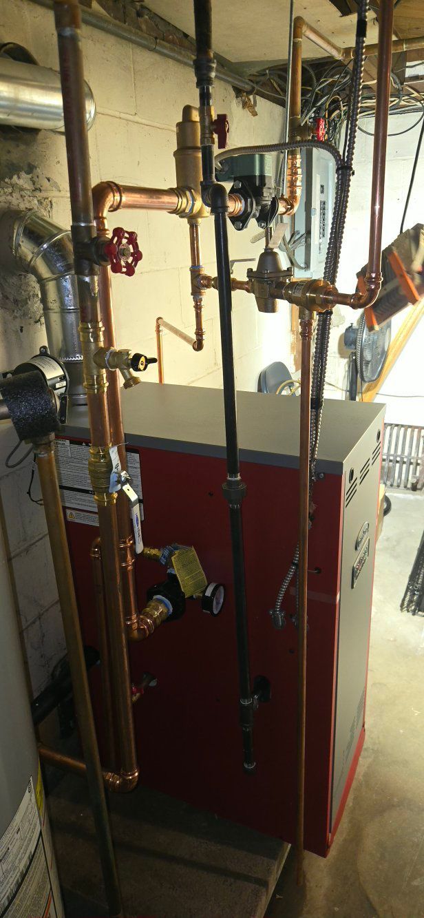A red and gray residential boiler unit installed in a basement, featuring extensive copper piping and plumbing valves.