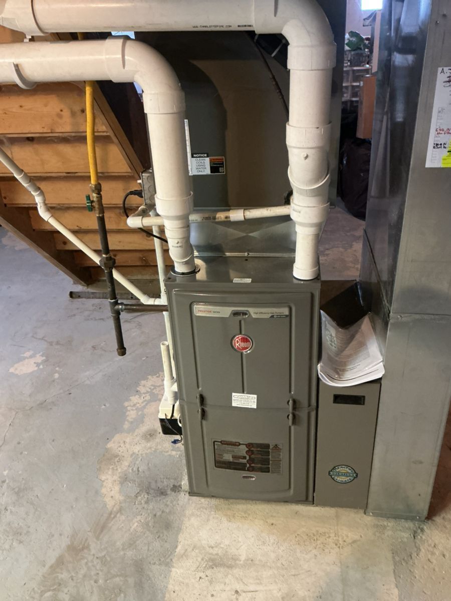 A grey residential furnace with connected white PVC pipes stands on a concrete floor in a basement.