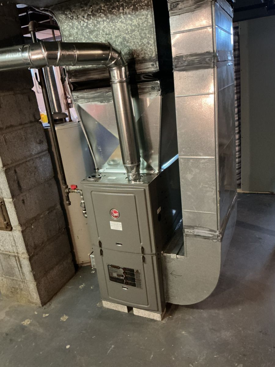 A grey residential furnace stands on blocks in a basement, connected to a vertical metal duct system and exhaust pipe.