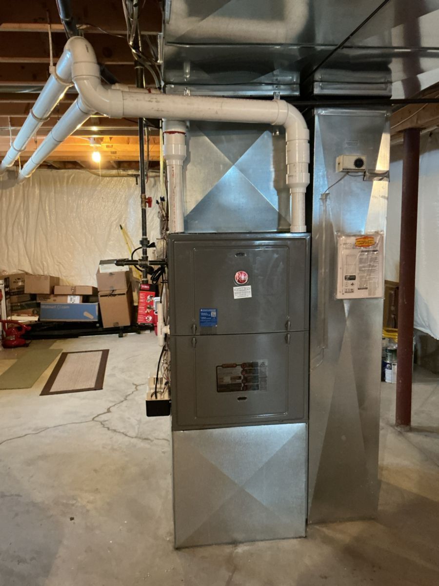 A grey residential furnace stands in a basement with attached white PVC exhaust pipes and galvanized steel ductwork.