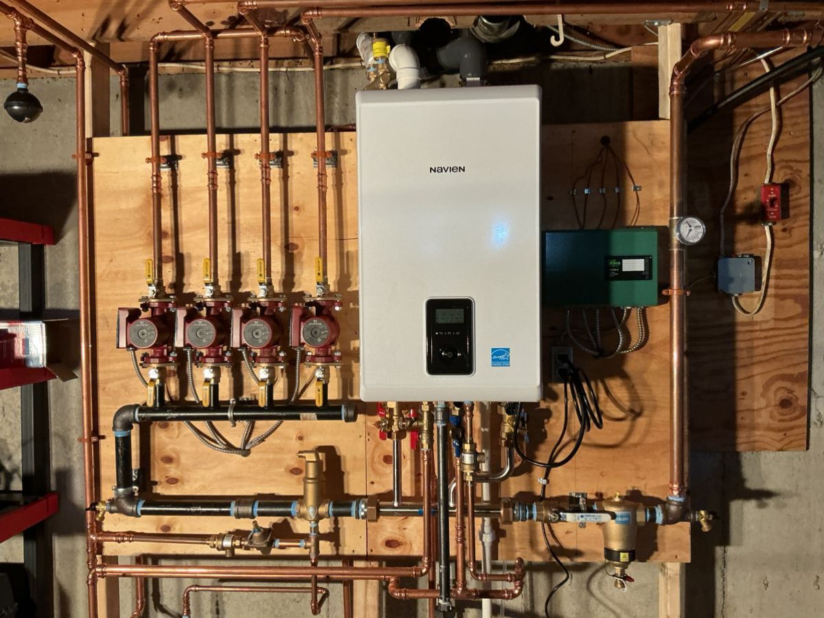 A white wall-mounted boiler unit with copper plumbing, pumps, and gauges mounted on a wooden backboard in a basement.