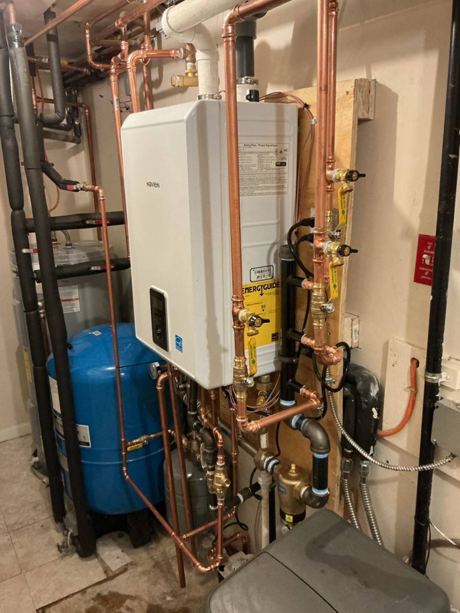 A white wall-mounted tankless water heater with extensive copper plumbing, valves, and a blue expansion tank in a basement.