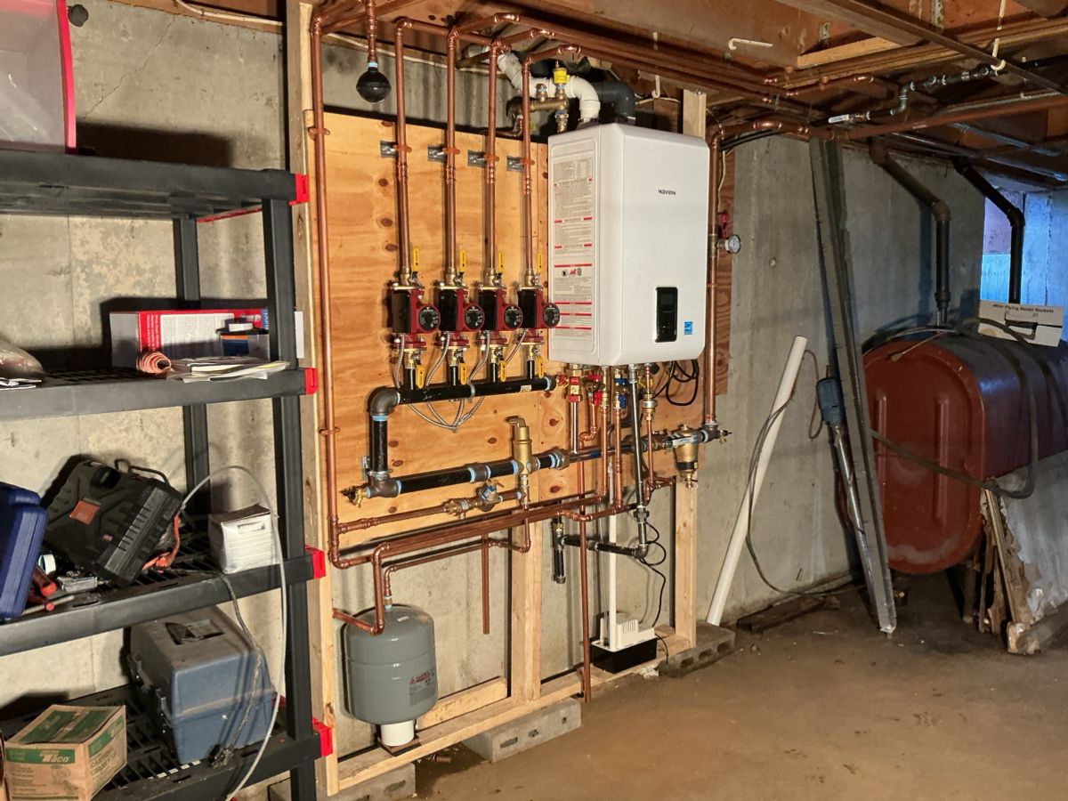 A white tankless water heater mounted on a wooden board with copper plumbing, pumps, and a grey expansion tank in a basement.