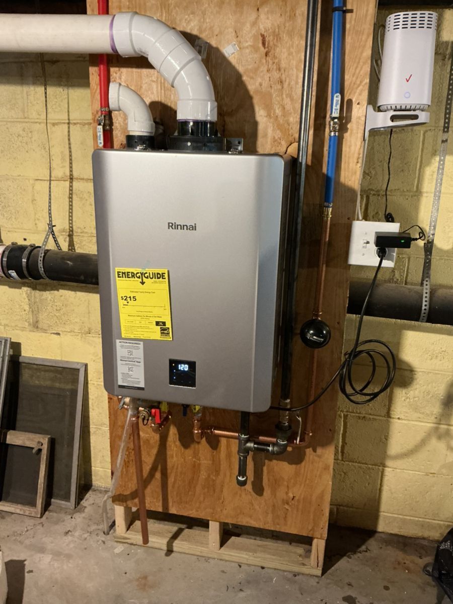 A Navien tankless water heater mounted on a wooden board in a basement, with connected white vent pipes and plumbing.