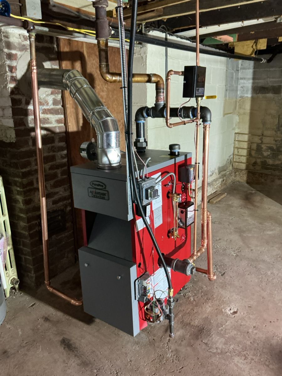 A red and gray boiler sits in a basement with connected copper and black iron piping against a brick wall.