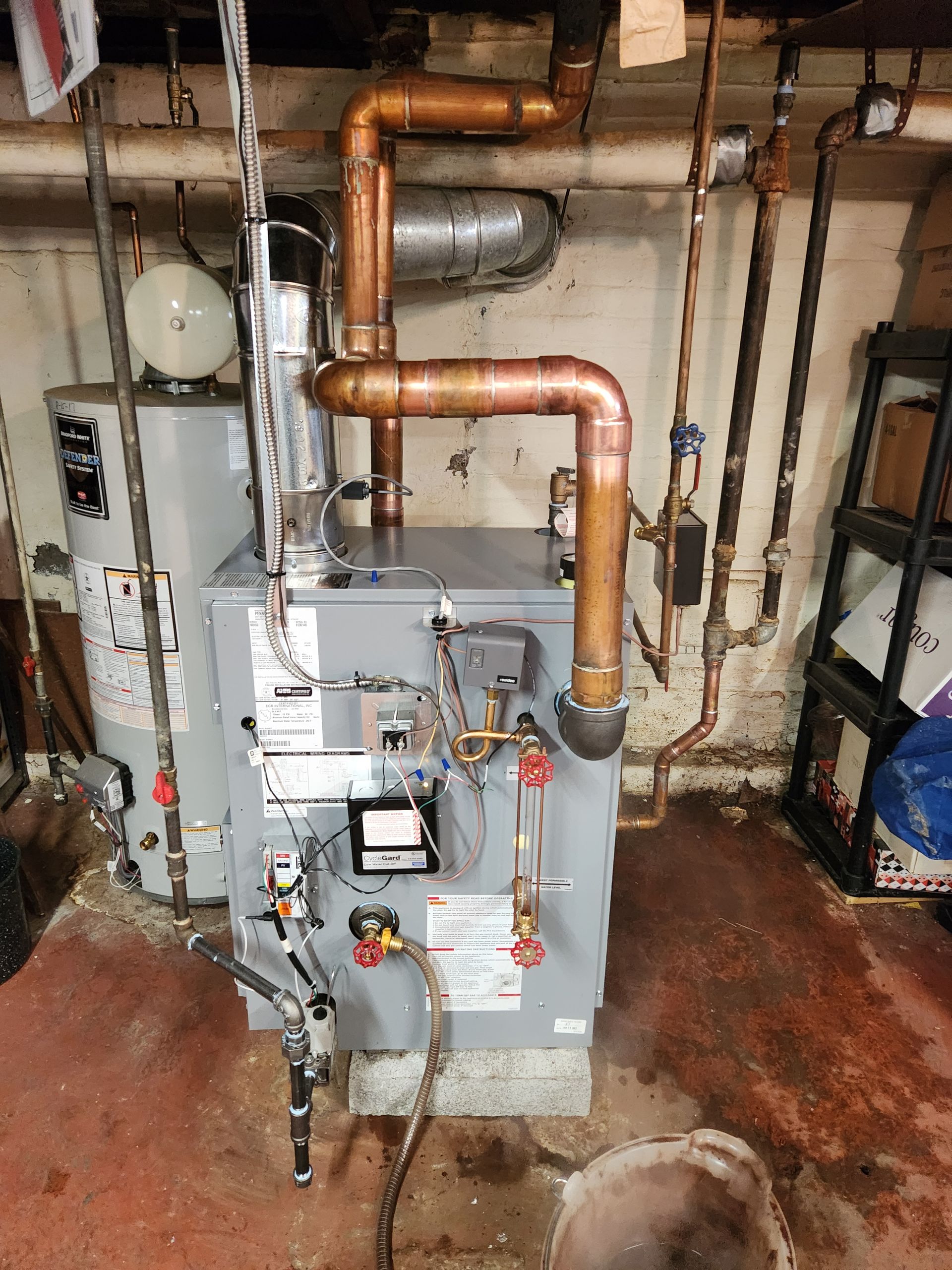 A gray residential boiler unit with copper piping and a metal vent pipe, situated in a basement setting.