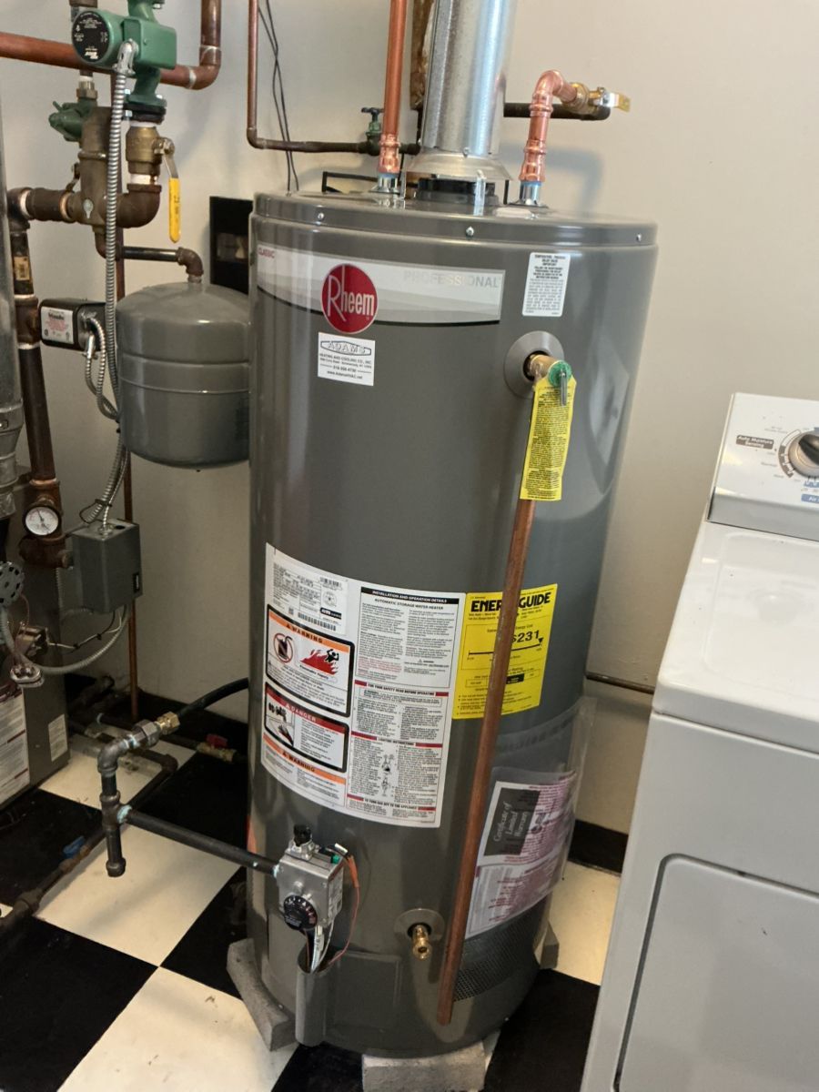 A tall, gray Rheem gas water heater stands in a room with a checkered floor, next to pipes and a laundry appliance.
