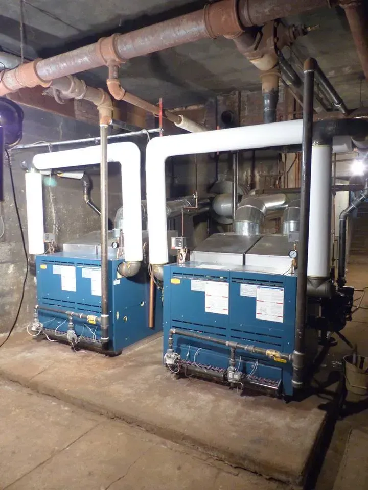Two industrial blue boilers with insulated white pipes stand on a concrete base in a basement.
