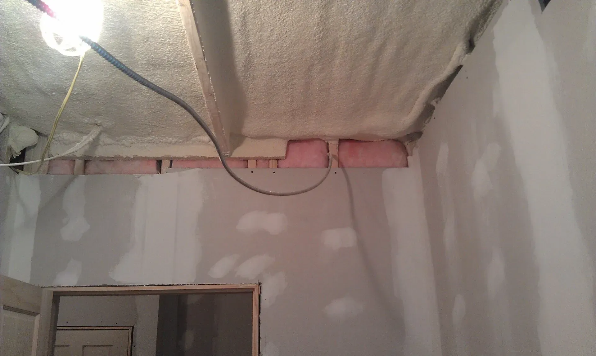 An unfinished room interior with spray foam ceiling insulation, pink fiberglass batts, and hanging electrical wires.