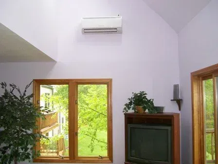 A white wall-mounted air conditioner sits high on a light purple wall above a window and a wooden TV stand.