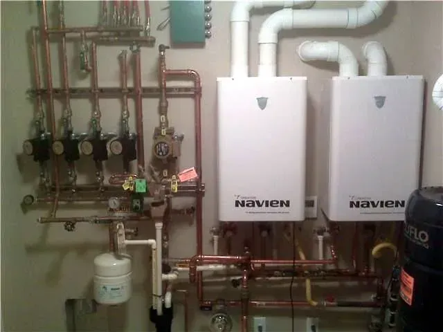 Two white Navien wall-mounted tankless water heaters connected to copper piping and a manifold system in a utility room.