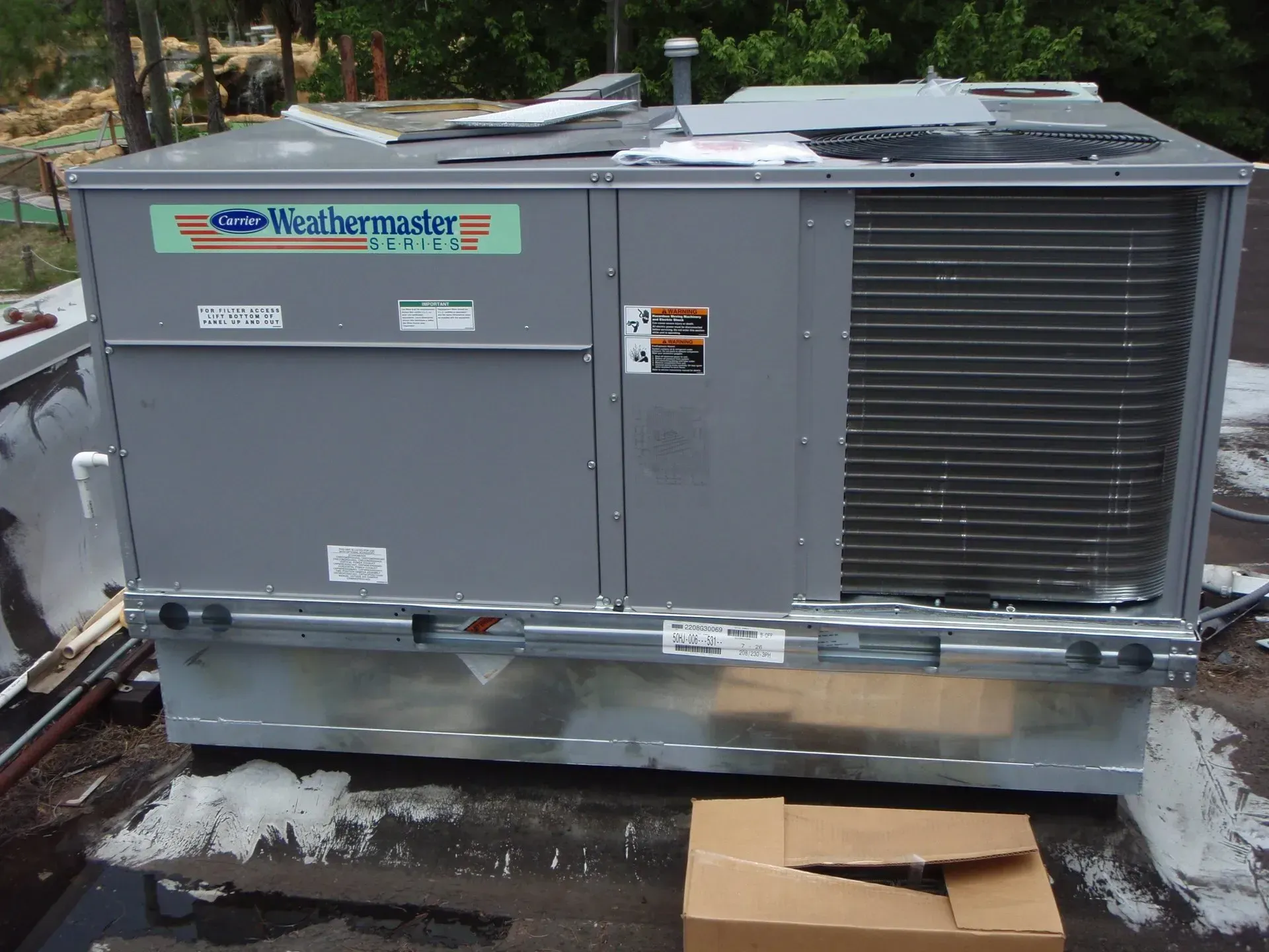A grey Carrier WeatherMaker rooftop HVAC unit sitting on a flat roof next to a cardboard box.