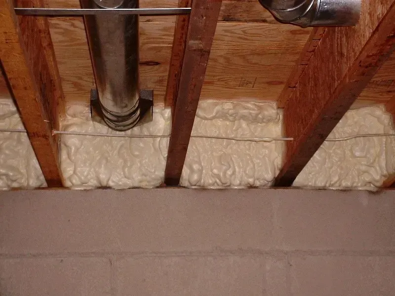 Spray foam insulation fills the rim joist cavities above a concrete basement wall, with a metal duct pipe visible.
