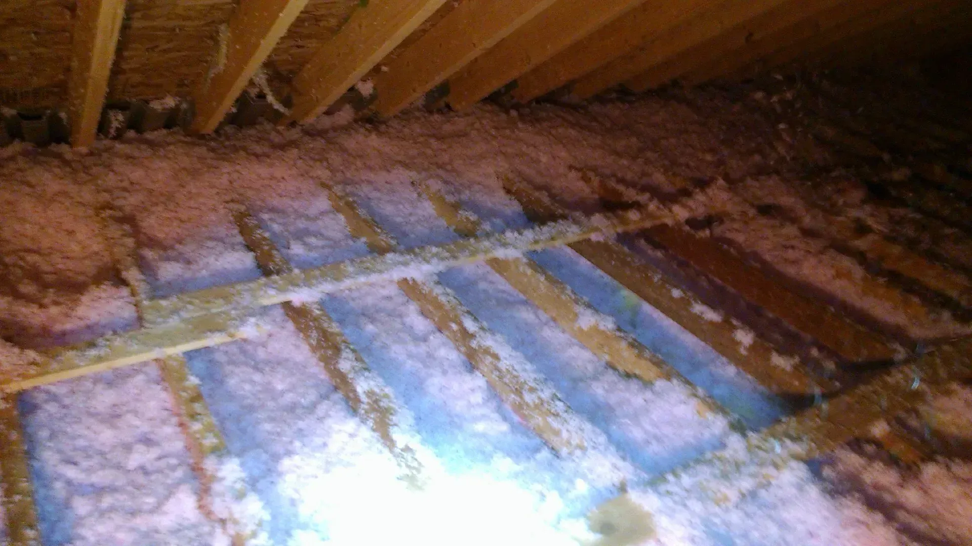 An attic floor showing mismatched pink and blue fiberglass insulation batts installed between wooden joists.