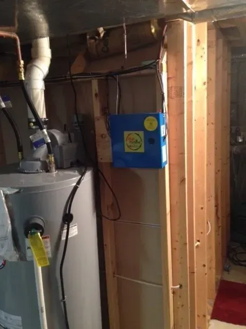 A water heater and a blue electronic air purifier mounted on a wooden wall in a utility room or basement.