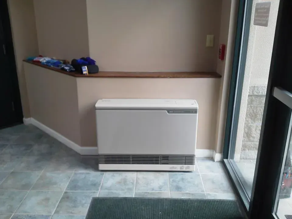 A white heater sits on the floor against a tan wall in an entryway with tiled flooring and a glass door.
