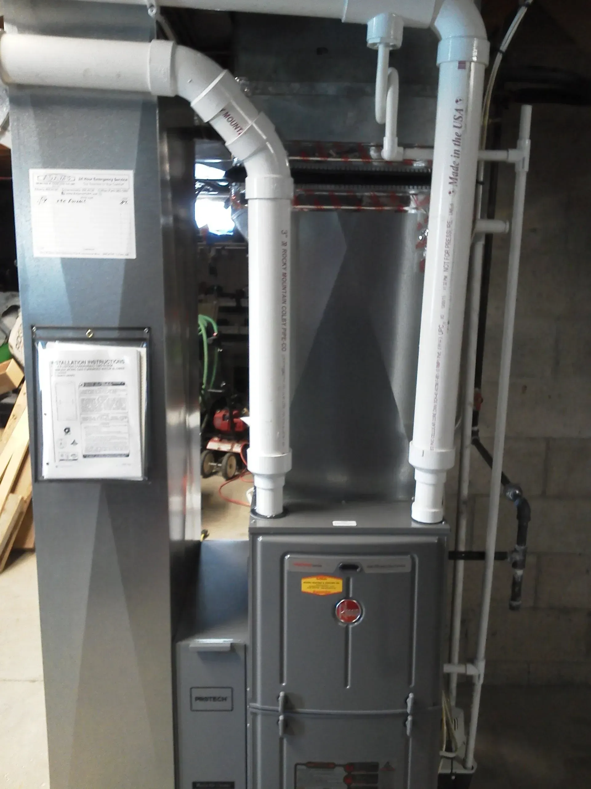 A grey HVAC unit in a basement with white PVC pipes attached to the top, against a cinder block wall.
