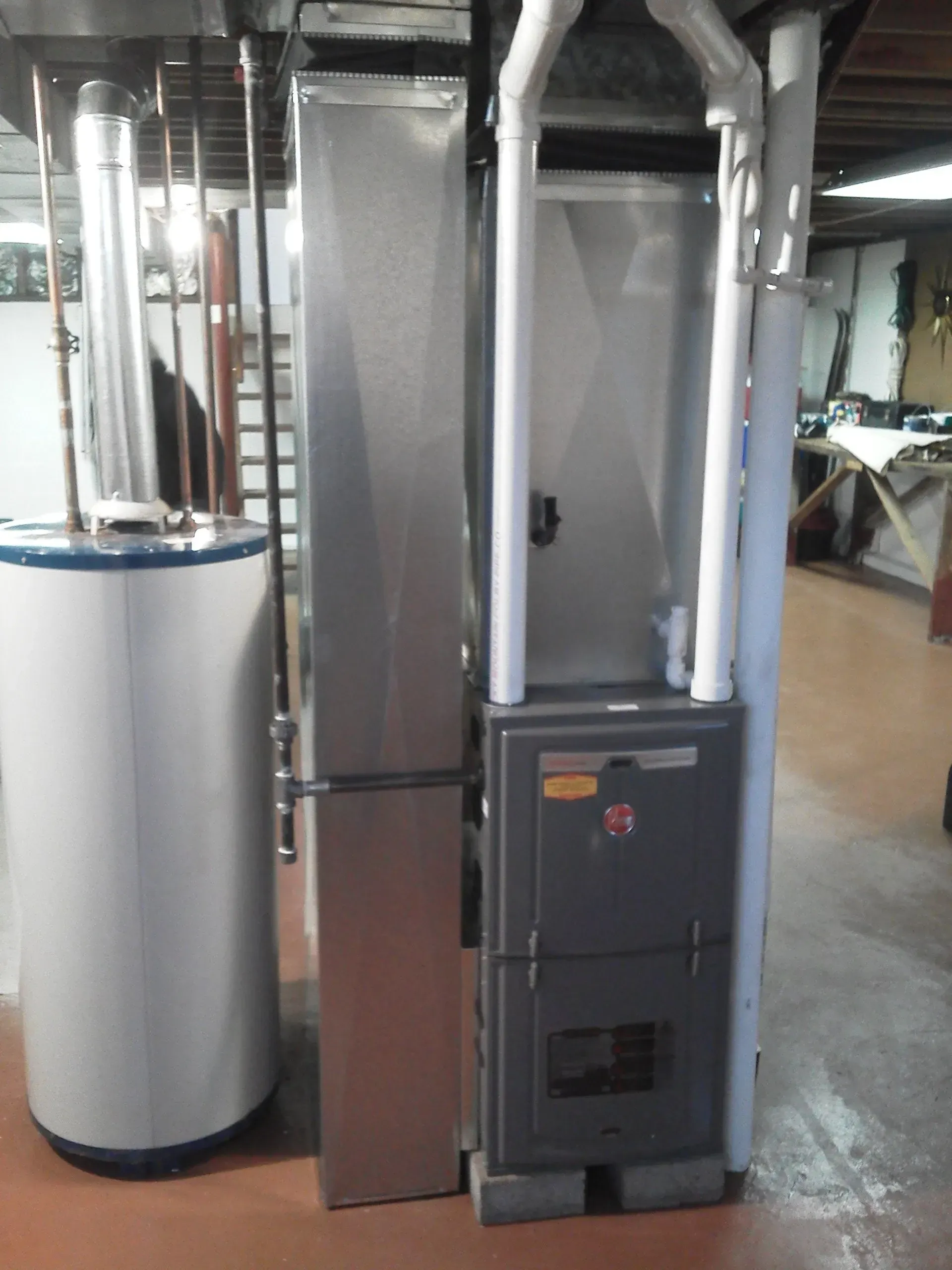 A water heater standing next to a grey high-efficiency gas furnace with white PVC venting pipes in a basement.