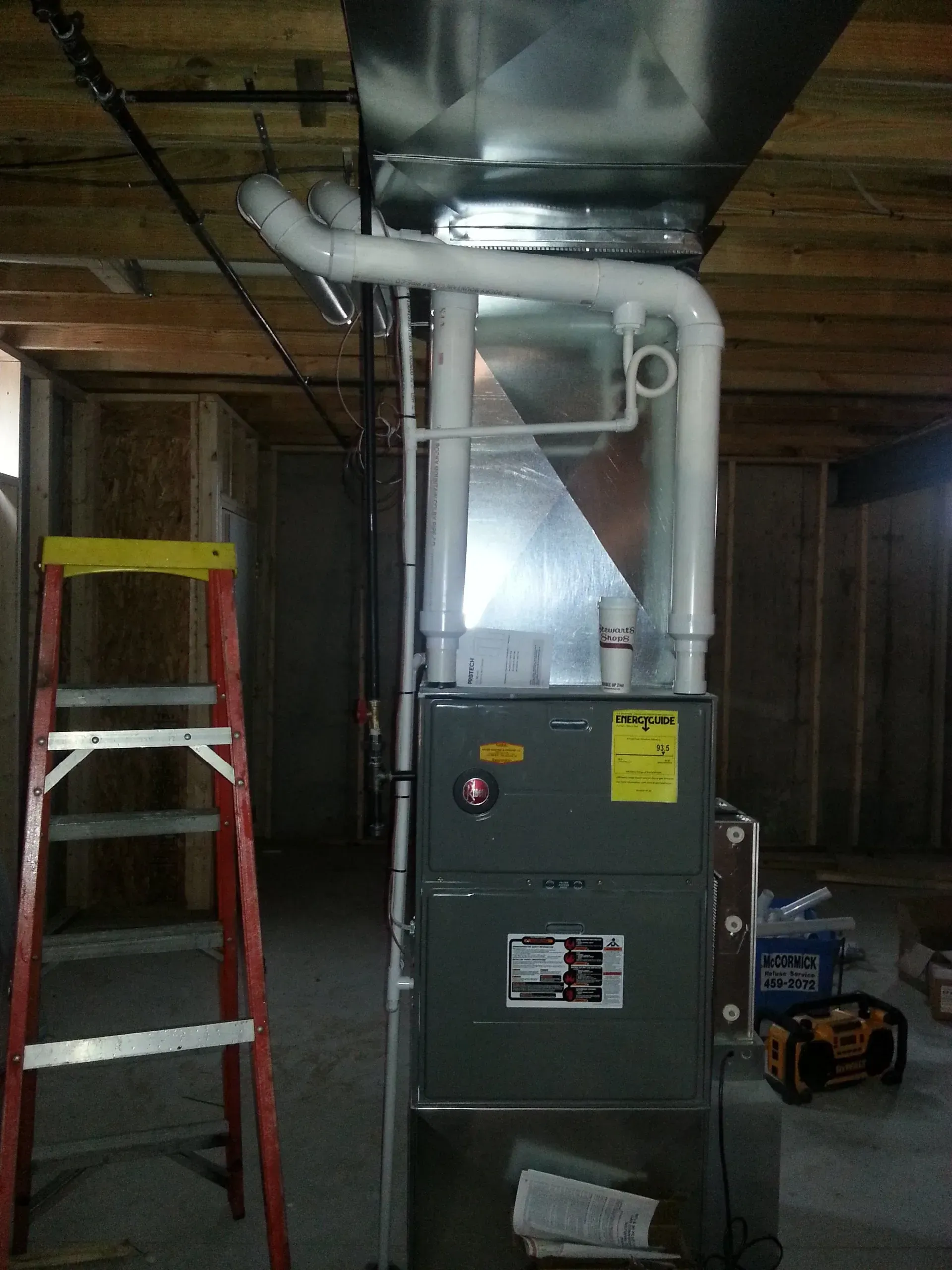 A grey gas furnace with white PVC venting pipes attached to the top, installed in an unfinished basement with a ladder.
