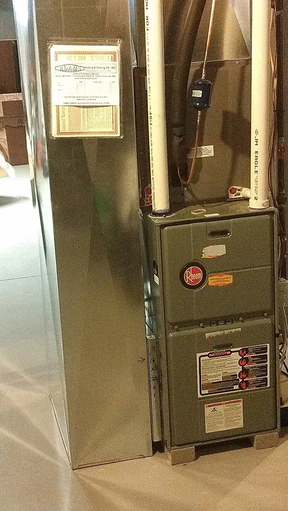 A green furnace unit sits next to a large, rectangular silver metal ductwork panel in a residential setting.