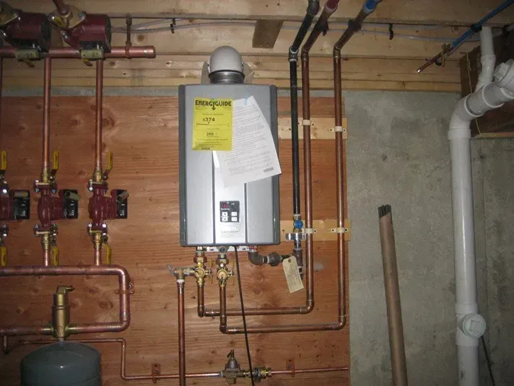 A tankless water heater mounted on a wooden wall with connected copper piping, valves, and pumps in a basement.