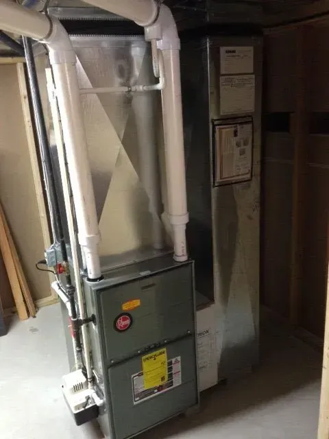 A high-efficiency gas furnace and AC coil unit with white PVC vent pipes installed in a utility room.