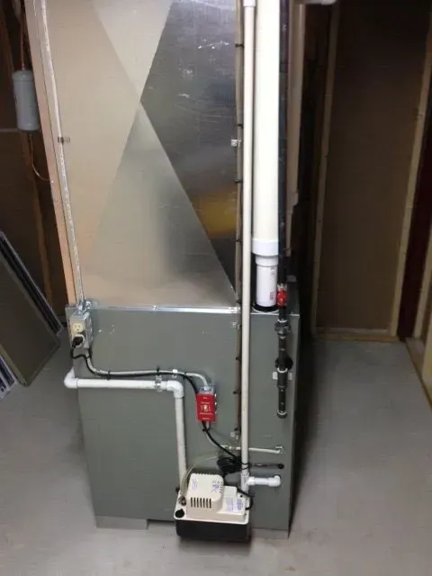 A residential HVAC furnace unit standing on a concrete floor with a white condensate pump and piping connected to the side.