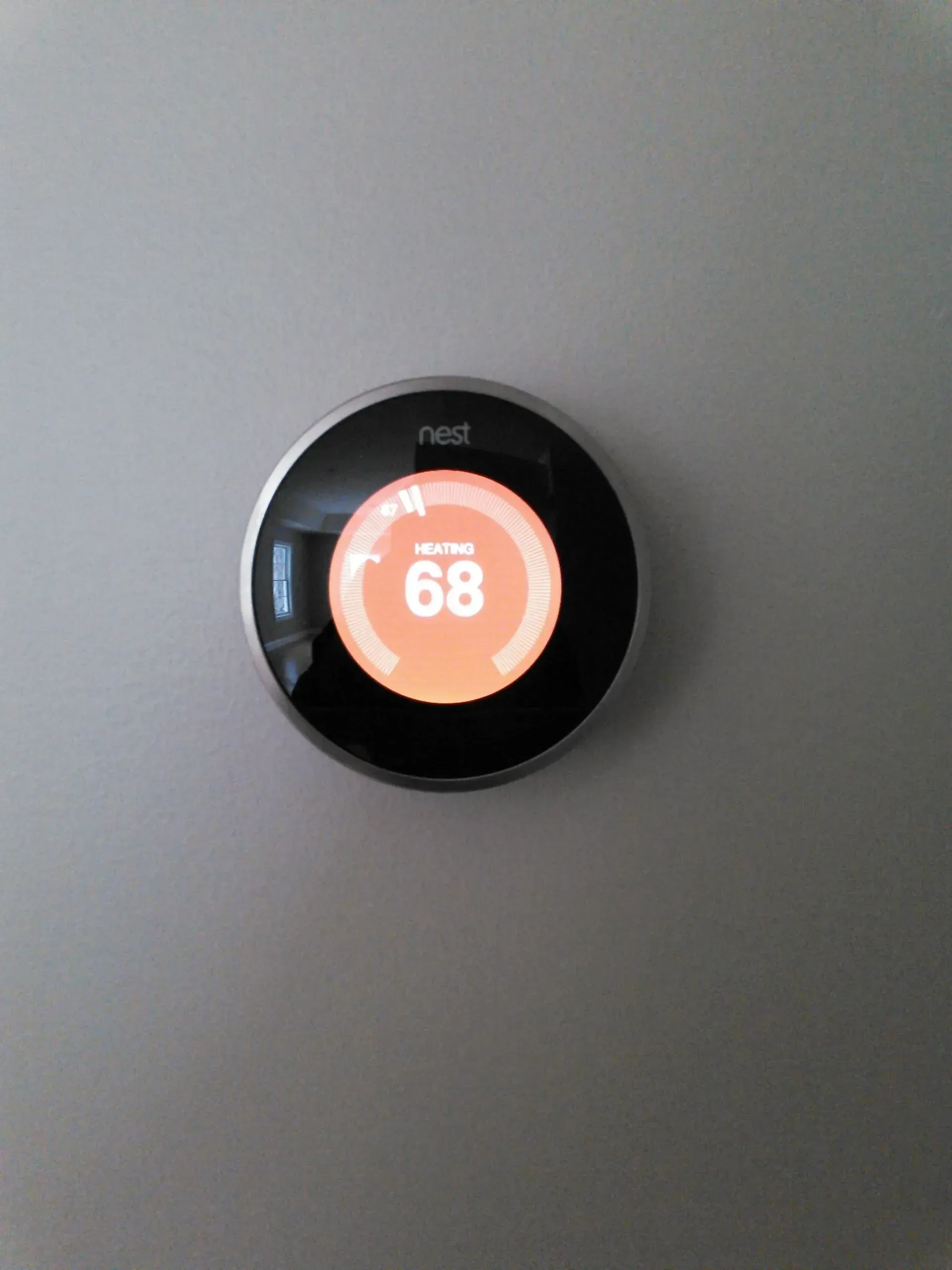 A Nest smart thermostat on a grey wall displaying an orange interface with a target temperature of 68 degrees.