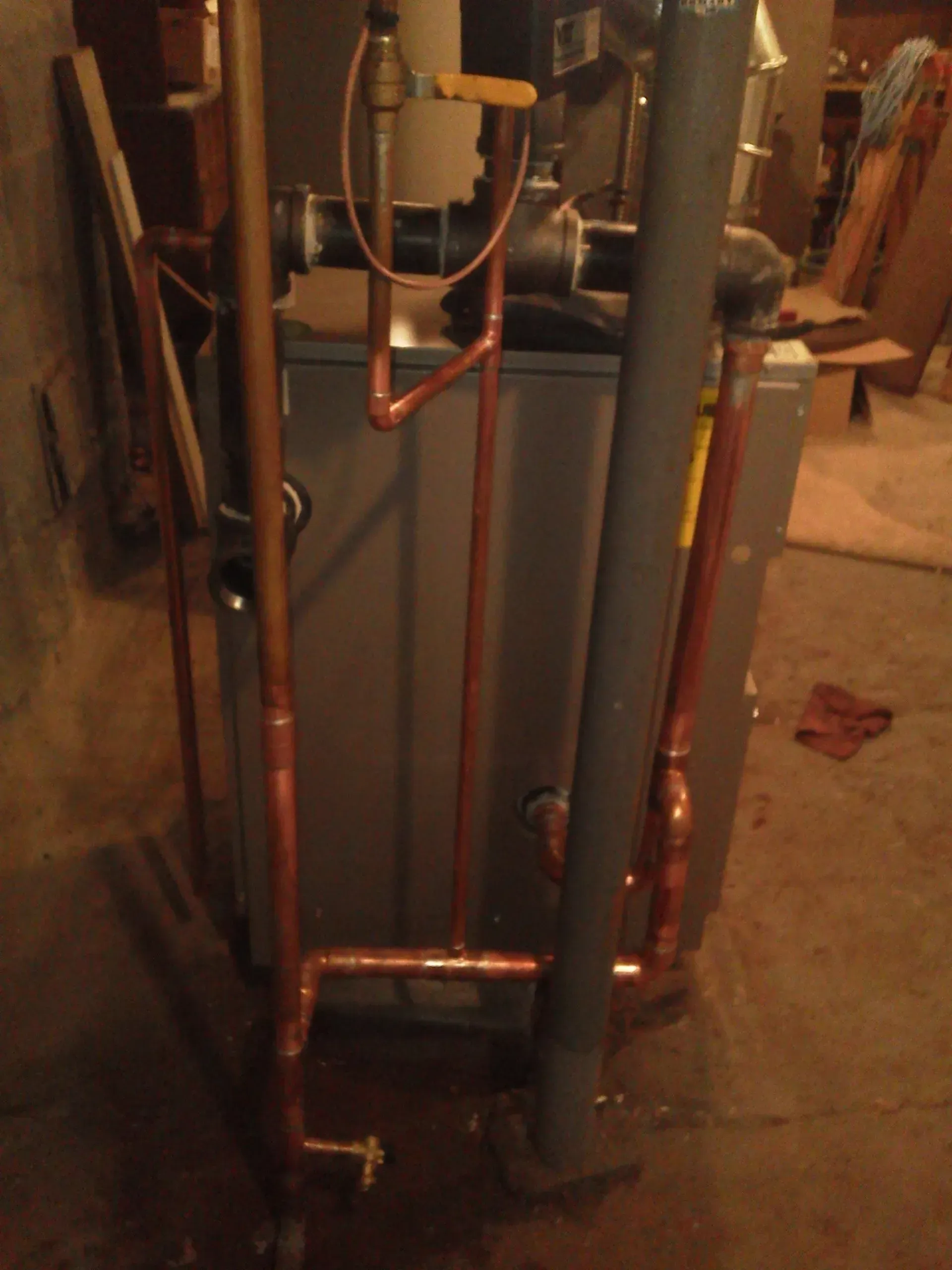 A central boiler unit in a basement, surrounded by copper plumbing pipes and a dark vertical exhaust pipe.