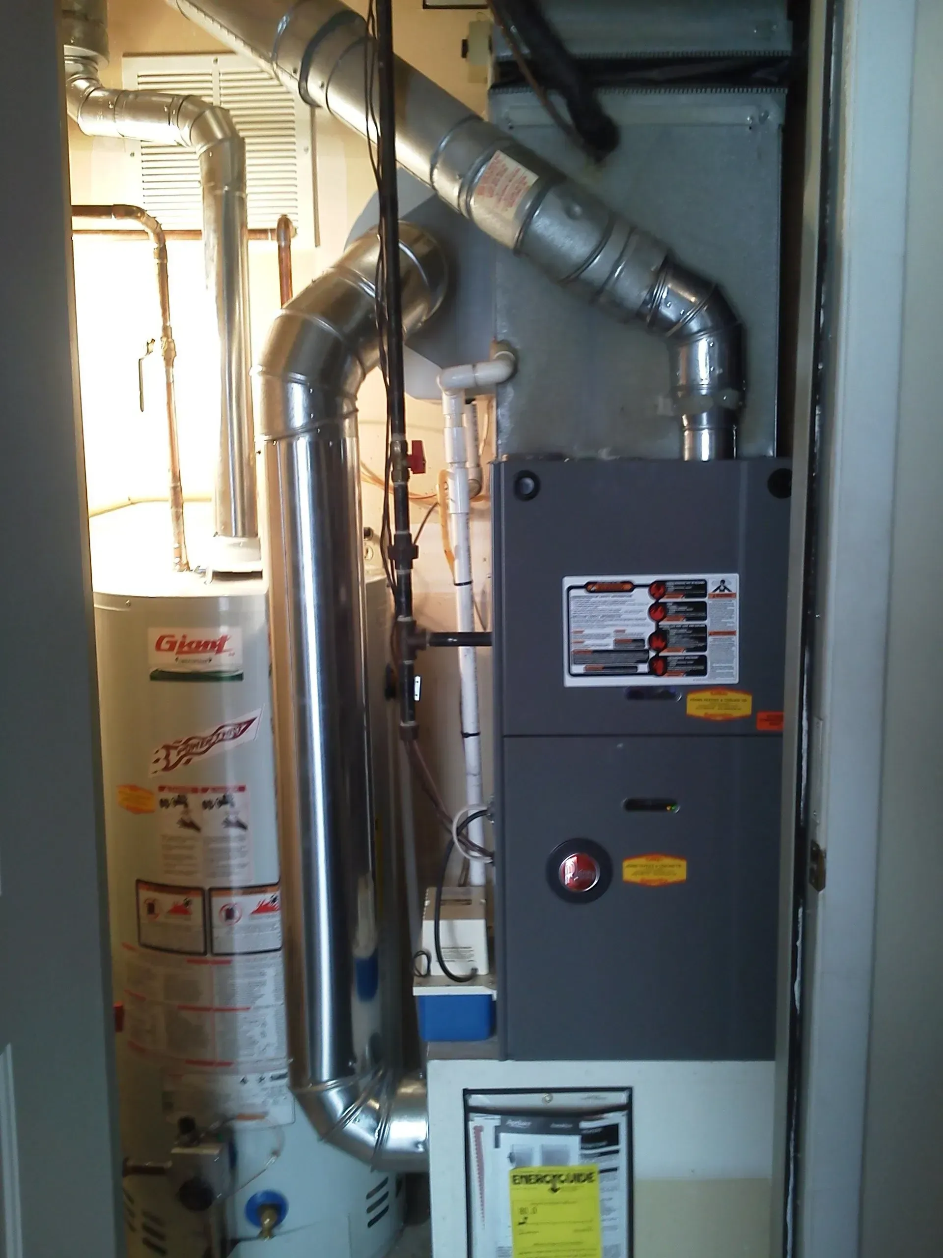 A utility closet containing a white gas water heater next to a grey furnace with attached silver metal ventilation pipes.