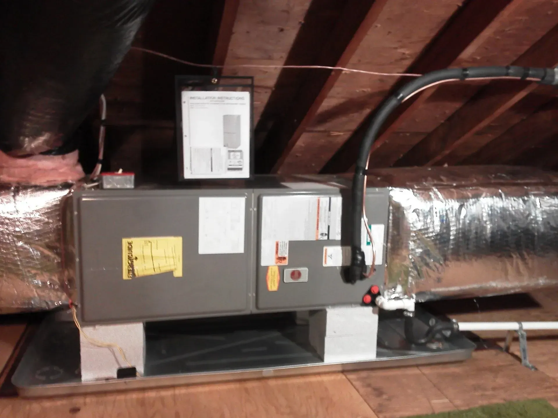 An attic HVAC unit mounted on cinder blocks inside a drip pan with connected silver ductwork and drainage pipes.