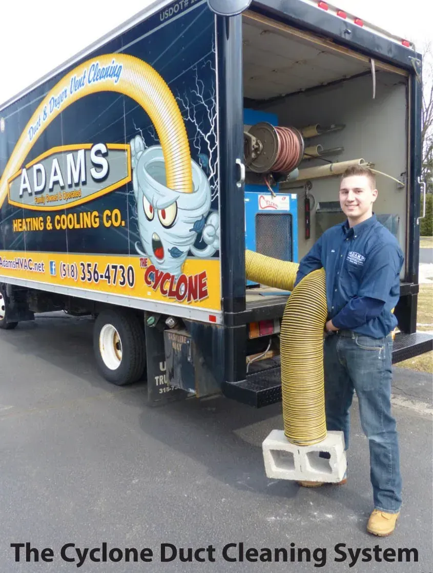 A worker stands beside an Adams Heating & Cooling truck, holding a large flexible yellow hose attached to a concrete block.