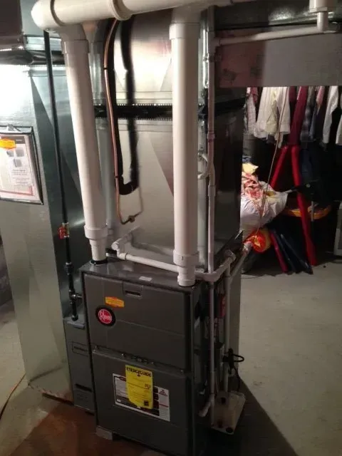 A grey high-efficiency gas furnace with white PVC intake and exhaust pipes in a residential basement setting.