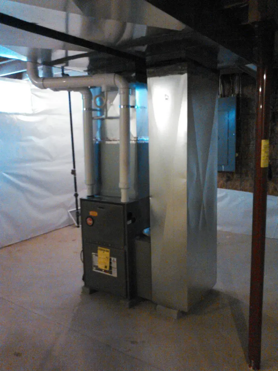 A modern furnace unit with white PVC vent pipes, standing in a basement with white wall liners and a metal support pole.