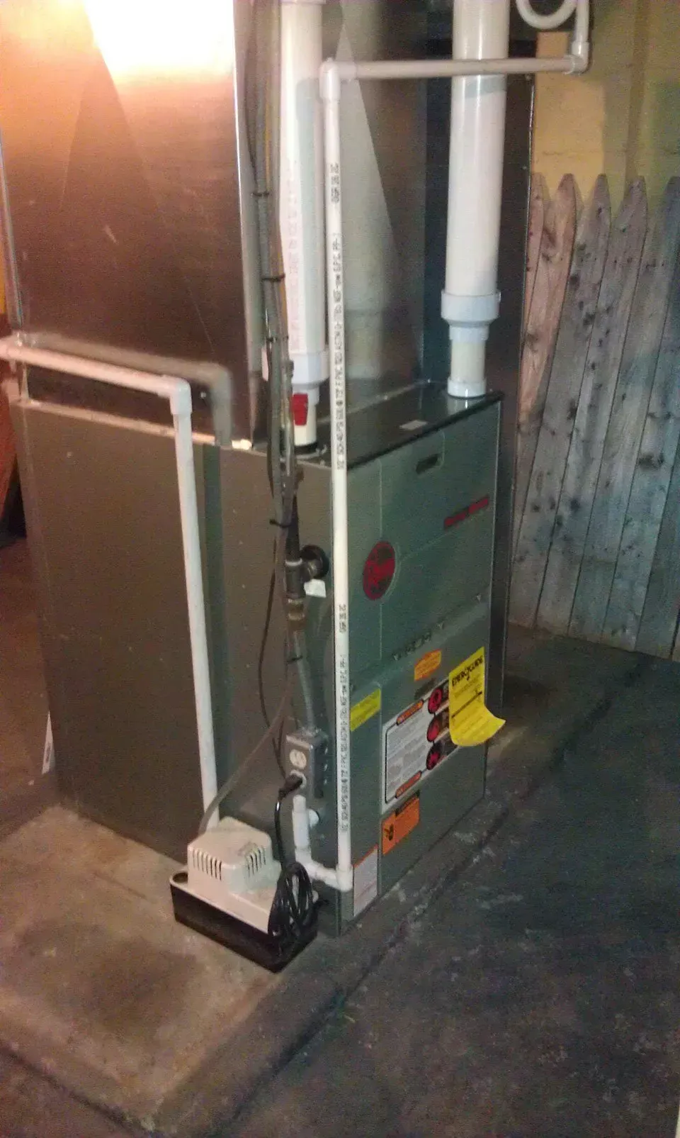 A grey high-efficiency gas furnace in a basement with white PVC intake and exhaust pipes and a condensate pump nearby.