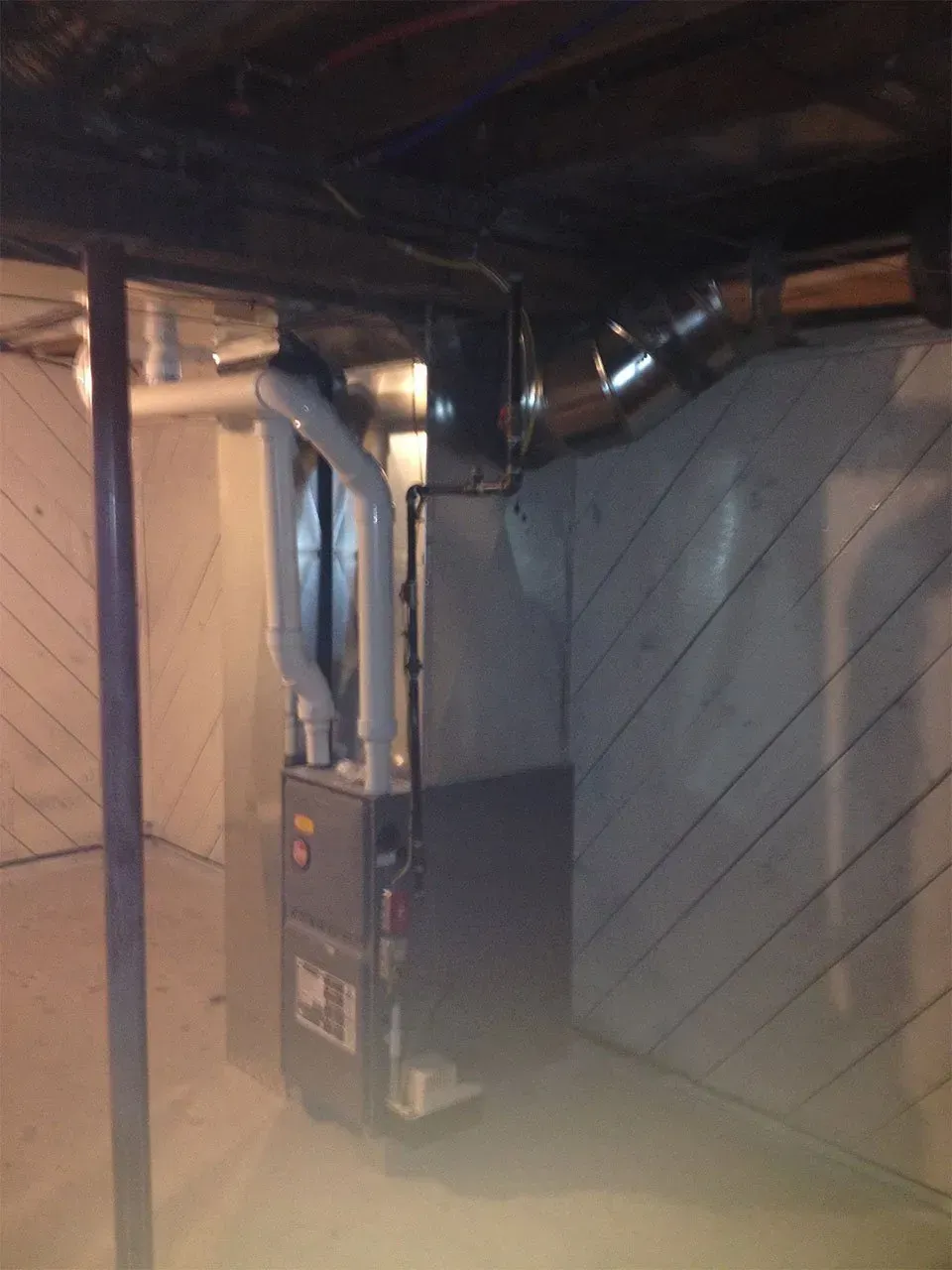 A gray high-efficiency furnace stands in a basement with connected white PVC venting and a metal ductwork transition.