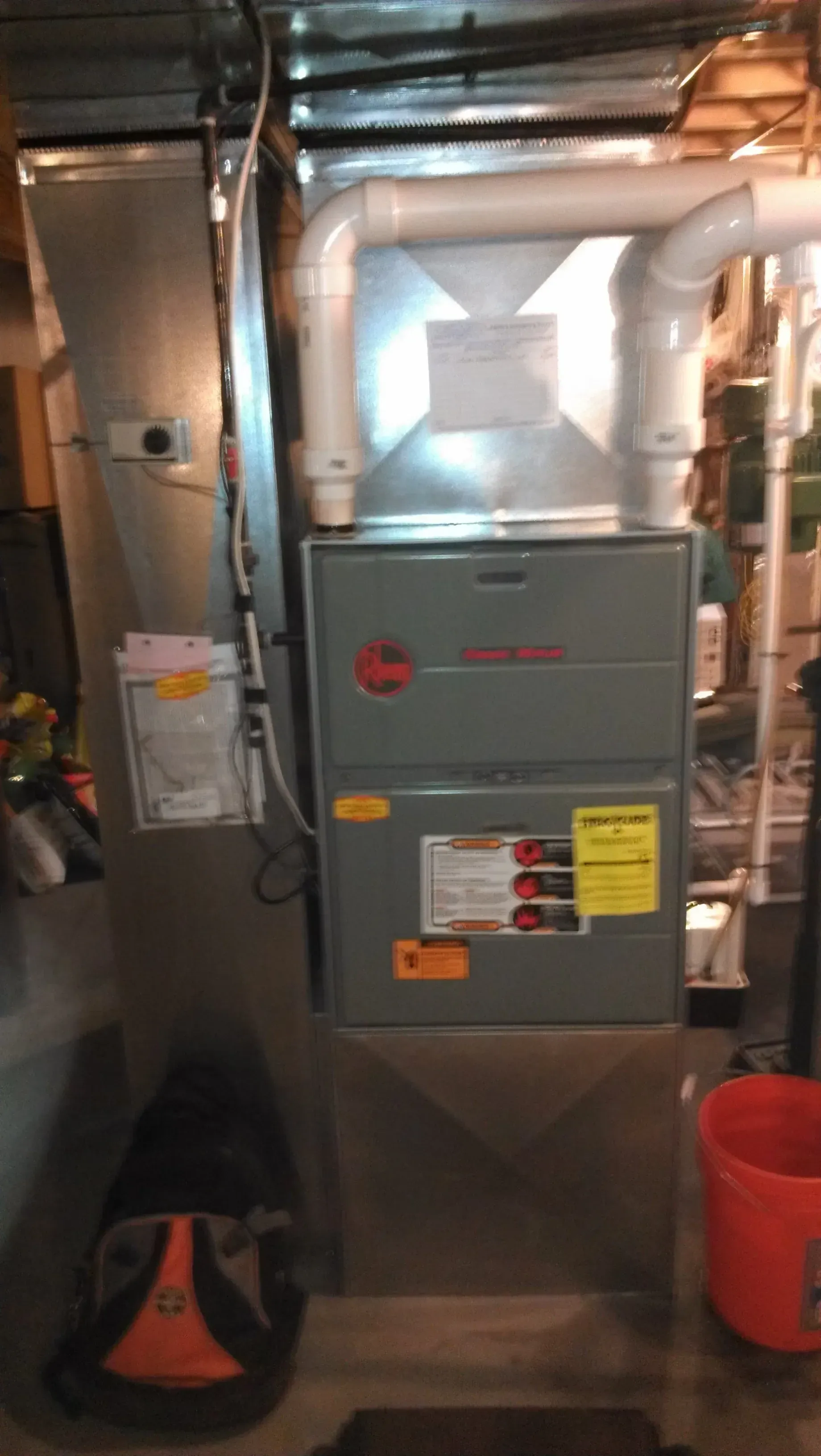 A gray Rheem furnace unit installed in a basement, featuring white PVC exhaust piping and metallic ductwork.