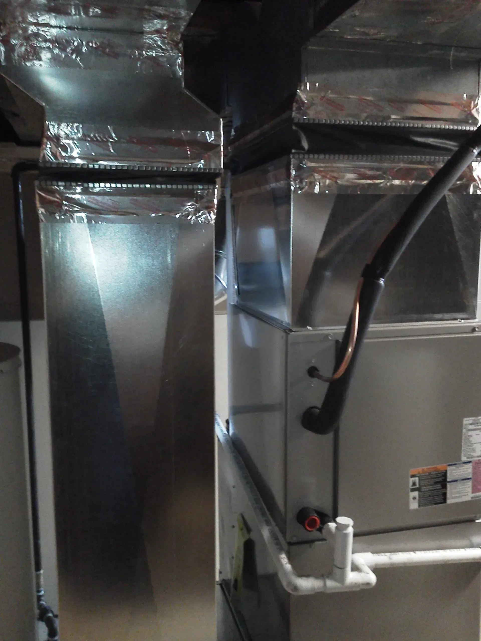 A vertical metallic HVAC air handler unit with connected ductwork and PVC plumbing pipes in a utility space.