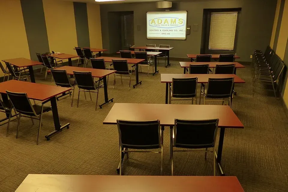 A classroom-style conference room with rows of tables and chairs facing a projector screen on a wall.