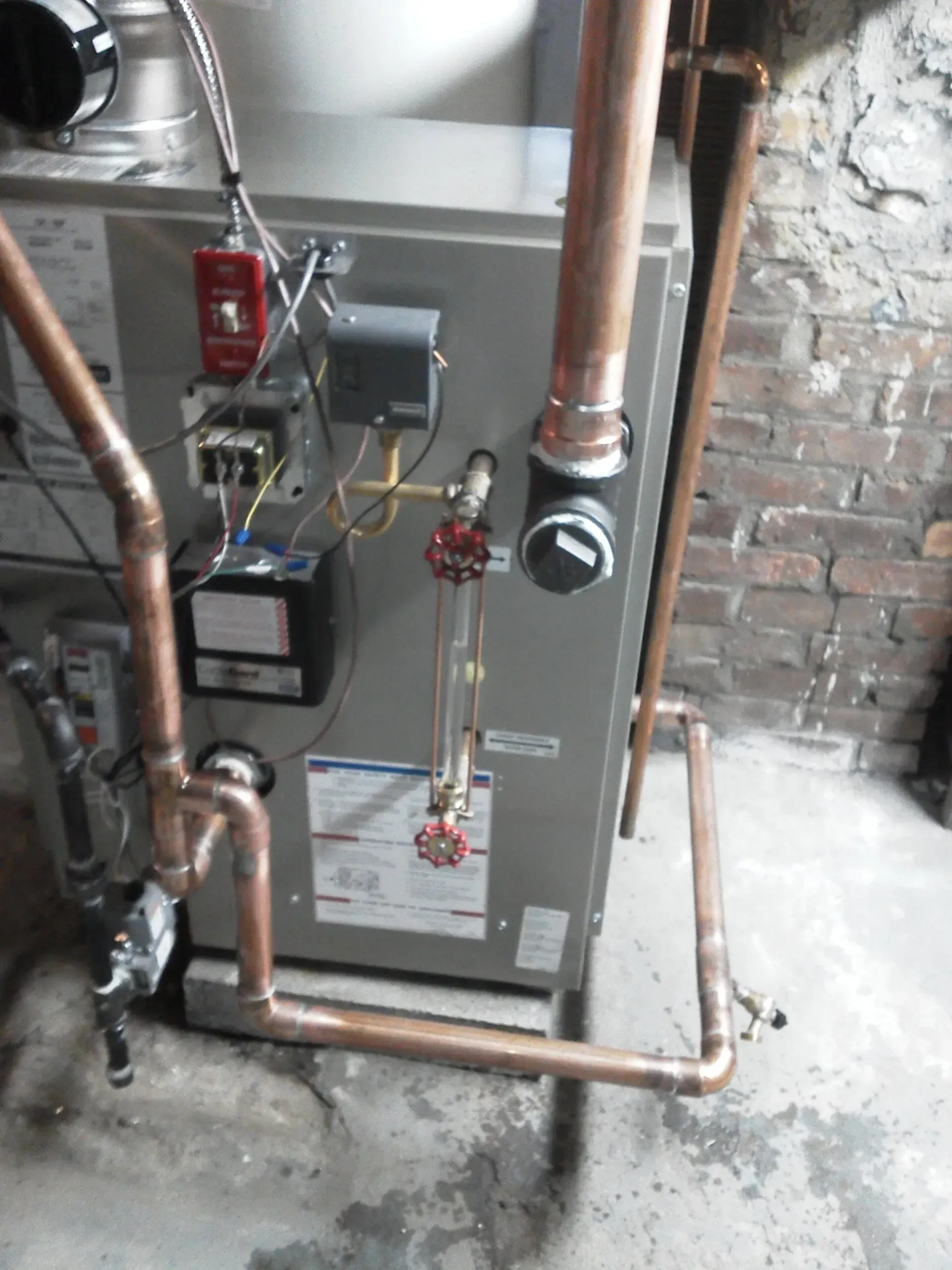 A gas-fired boiler with connected copper plumbing and a glass water level gauge installed against a brick wall.
