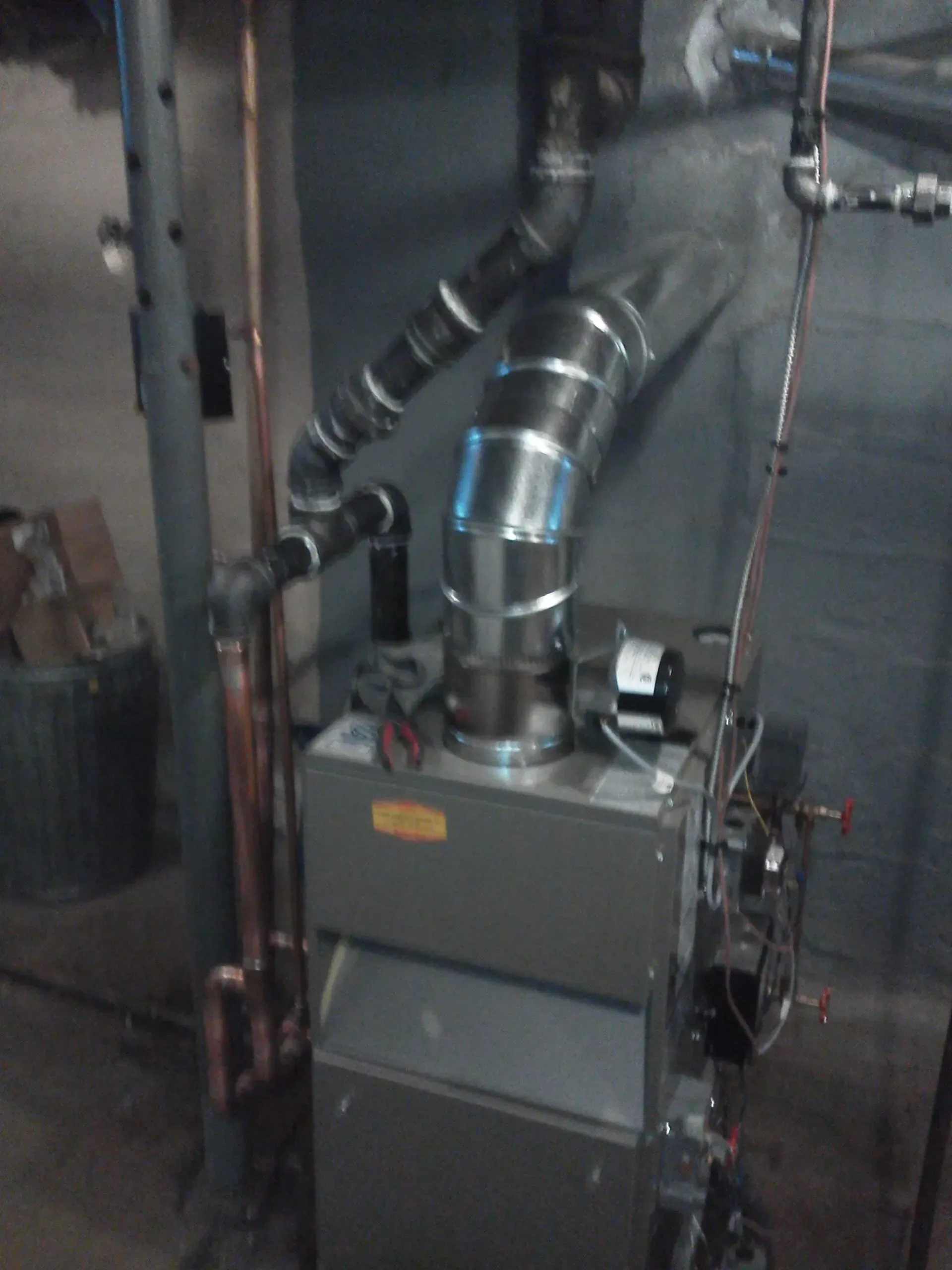 A grey boiler unit in a basement, featuring vertical silver venting and copper piping connections.