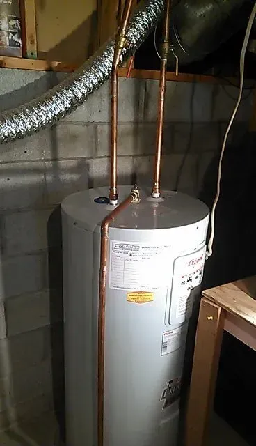 A light-gray water heater with two vertical copper pipes on top, situated in a basement against a concrete block wall.